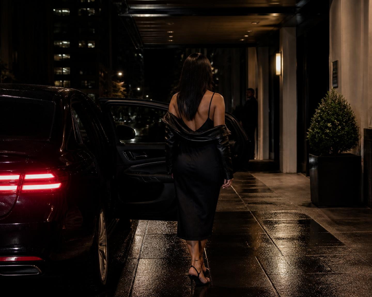 A woman in a black slip dress with a leather jacket over her shoulders, seen from behind, stepping from a black sedan toward a hotel entrance at night.